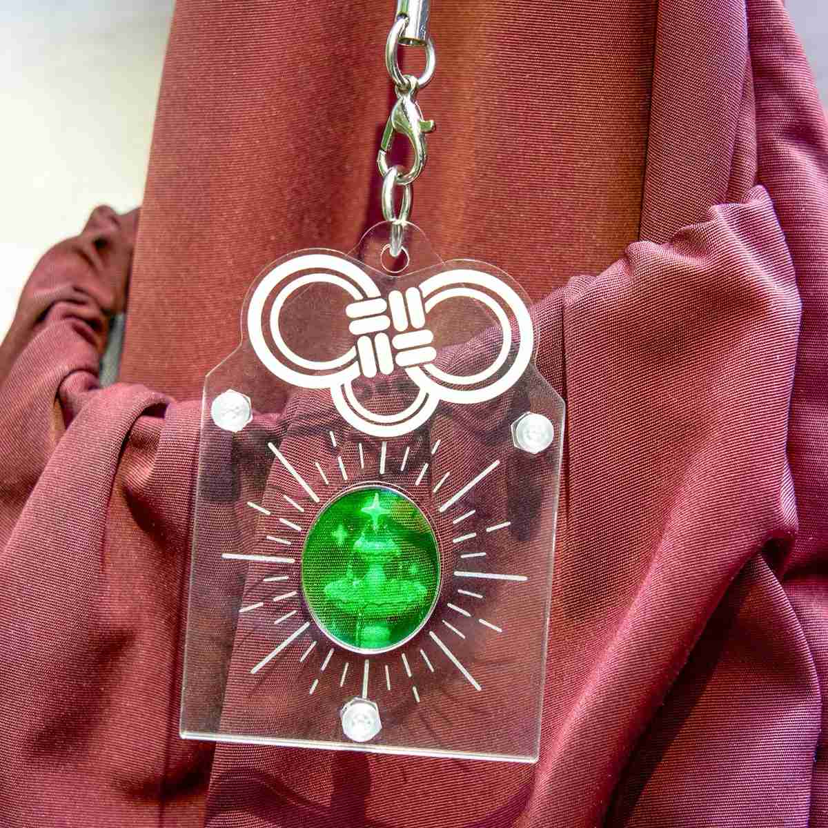 A clear acrylic keychain featuring a white printed knot design at the top and a green circular pendant with a 3d hologram wishing well illustration in the center, displayed against a draped burgundy fabric background.
